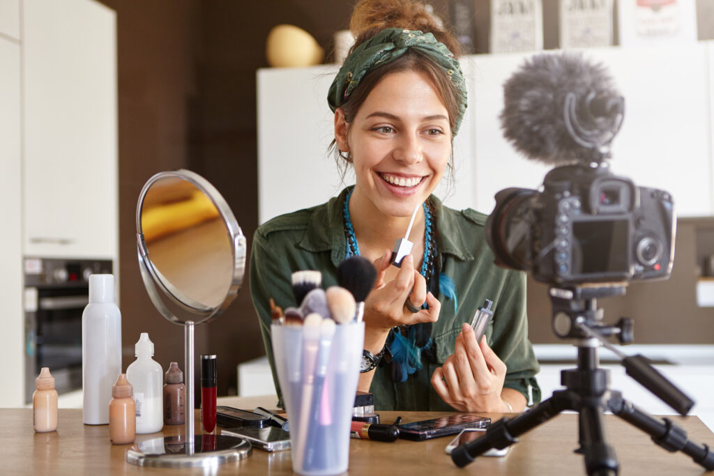 Beauty, fashion, technology, media and networking. Pretty girl vlogger sitting at dressing table surrounded with diverse cosmetics, looking at camera fixed on tripod while recording make up tutorial